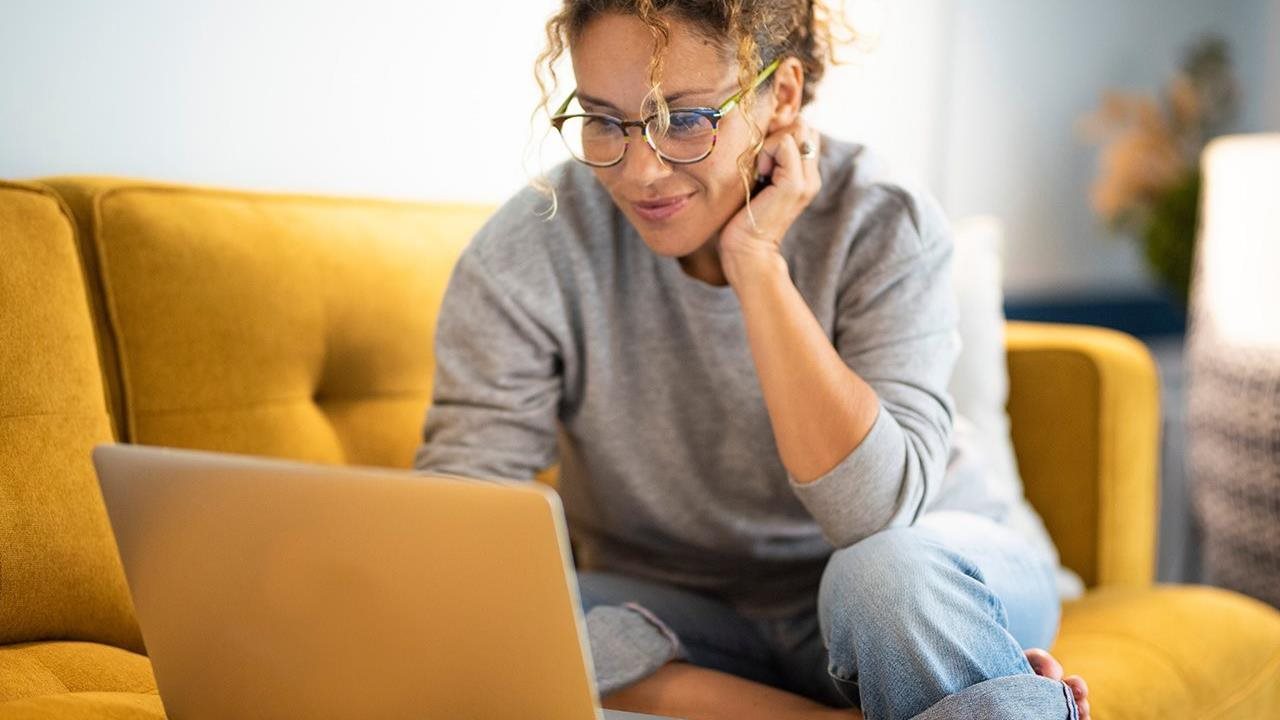 A woman with her curly hair in a bun rests her head on her palm and smiles as she looks at her laptop.