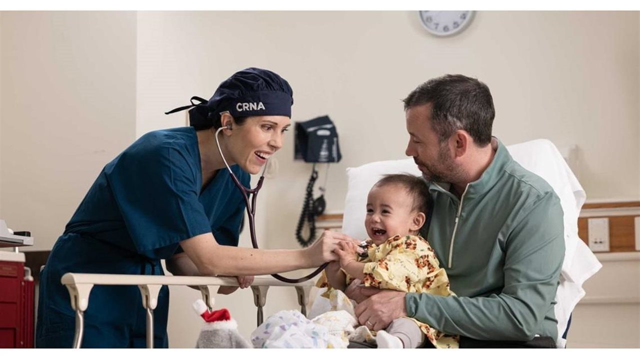 Nurse listens to baby's heart while dad holds him close. AANA represents Certified Registered Nurse Anesthetists (CRNAs)