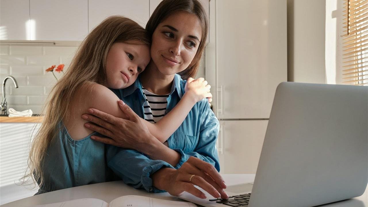 Woman working on a laptop computer at home while her daughter gives her a hug.