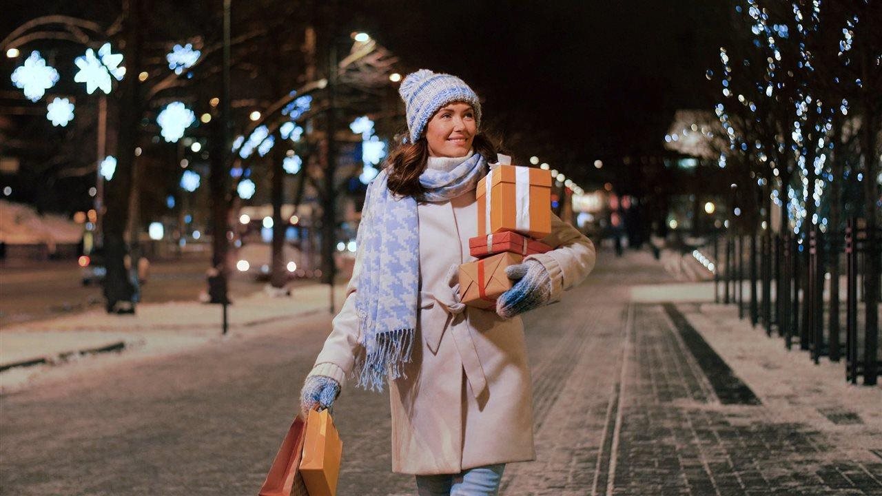 Woman wearing a white coat with blue winter hat and scarf wallking on a downtown street carrying holiday gift boxes.